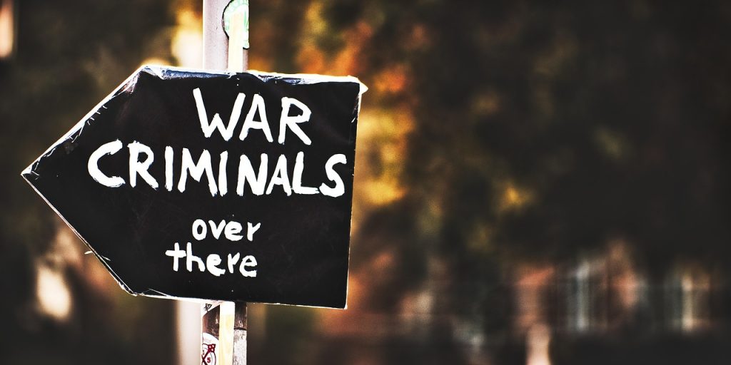 | What Happens When Fear Becomes Policy A handmade black sign shaped like an arrow is attached to a pole. The sign reads “WAR CRIMINALS over there” in bold white letters, pointing to the right. The background is blurred, with warm autumn tones and soft light. The image conveys protest, accusation, and political urgency.