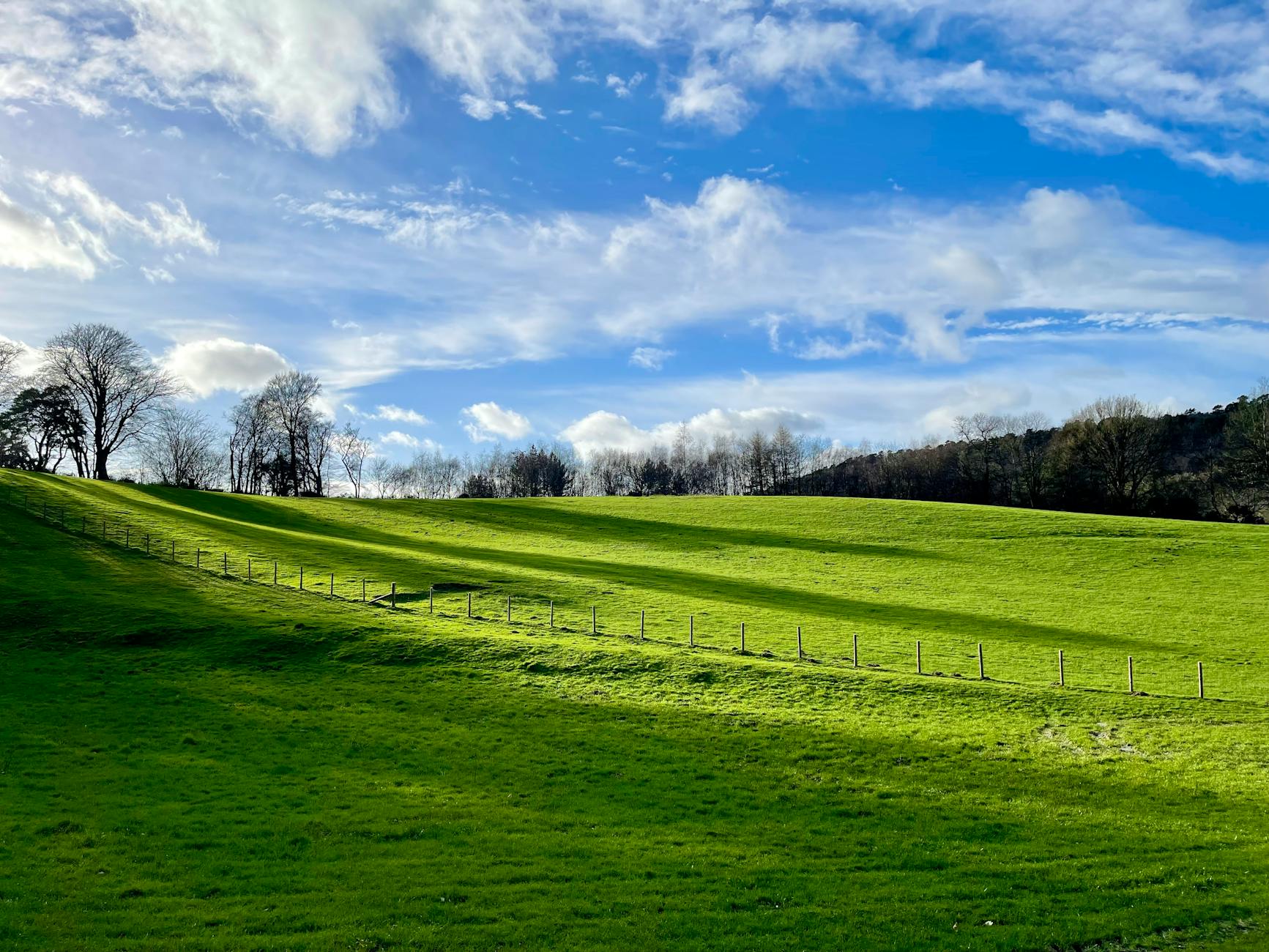 Rolling green countryside with a fence line across grassy hills under a blue sky with scattered clouds.